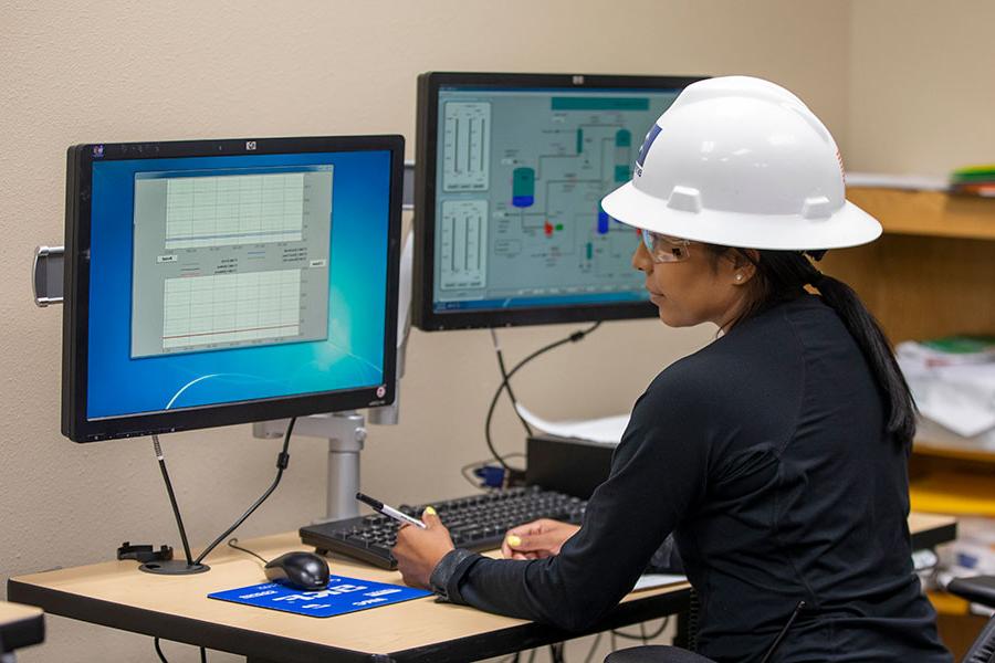 An individual in a white hard hat and safety goggles sits at a desk looking at a computer screen